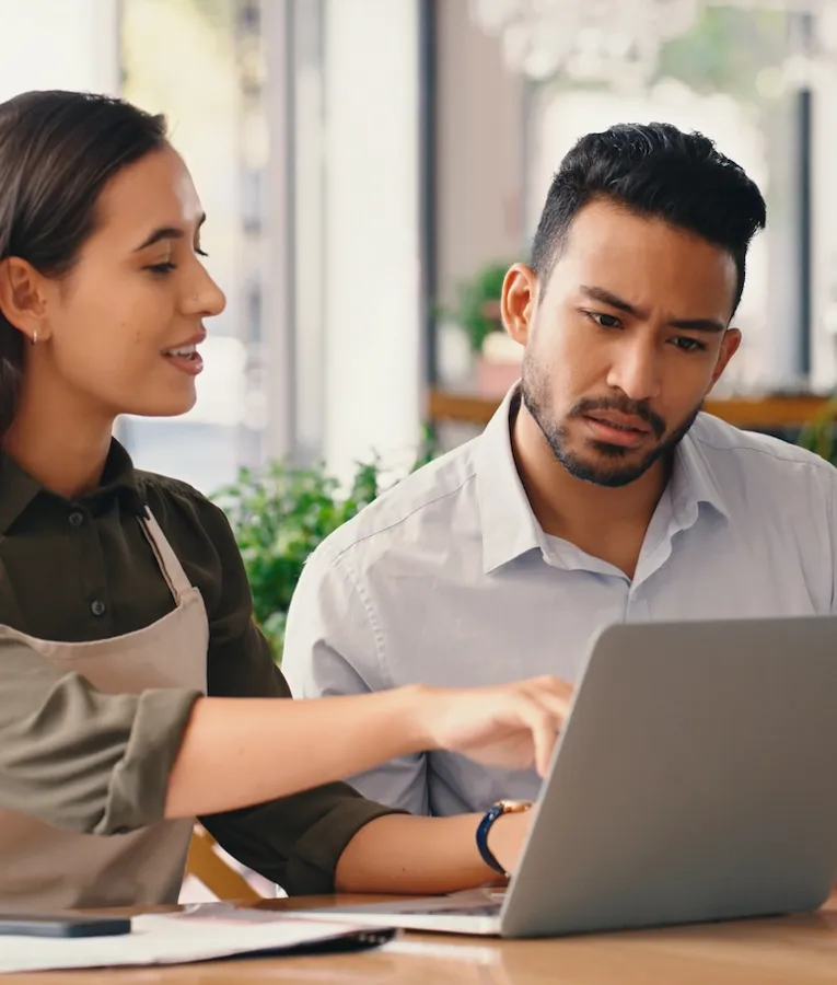 En kafémedarbeider i forkle og en eier gjennomgår økonomiske dokumenter sammen på en bærbar PC i en moderne kafé.