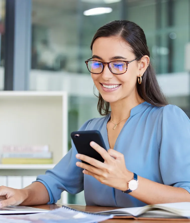 Smilende kontoransatt i blå bluse bruker smarttelefon og bærbar PC ved skrivebord – illustrerer digital arbeidsflyt, fjernarbeid og effektivitet på moderne arbeidsplass.