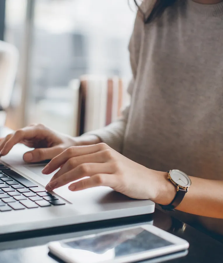 En fjernarbeider i en beige genser skriver på en bærbar PC i et moderne arbeidsmiljø. En smarttelefon ligger på det mørke trebordet, som symboliserer en fleksibel og digital arbeidshverdag.