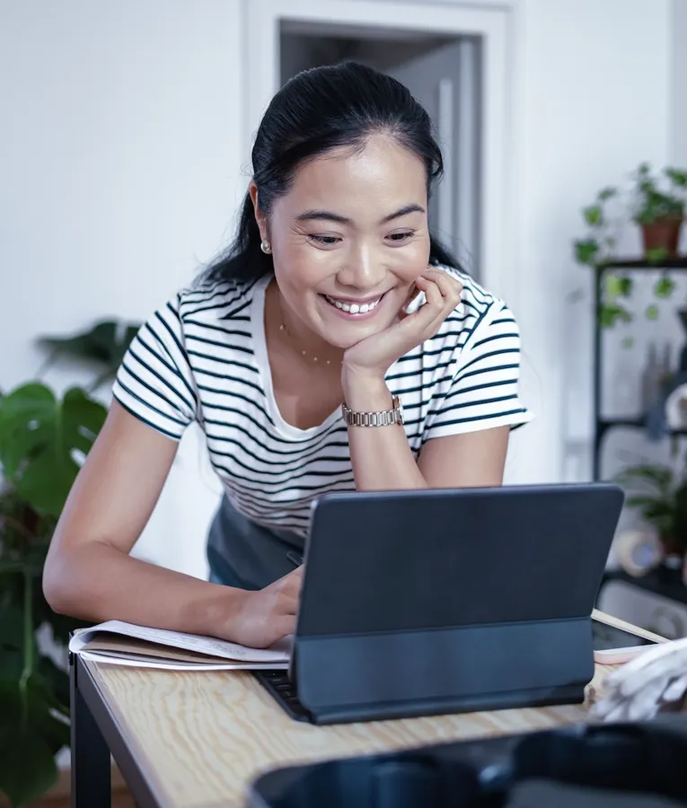 En smilende profesjonell i en stripete topp lener seg over et trebord mens hun jobber på et nettbrett med tastatur og deltar i en produktiv samtale på arbeidsplassen.