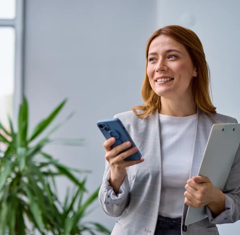 En smilende forretningskvinne i en grårutete blazer holder en smarttelefon og en skriveplate mens hun står i et lyst, moderne kontor med en grønn plante i bakgrunnen.