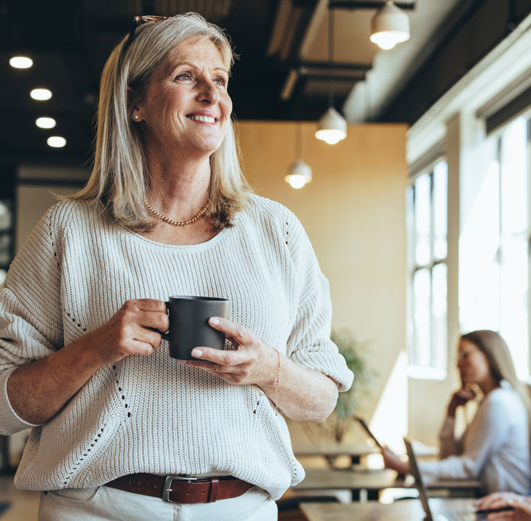 En smilende eldre kvinne med grått hår, iført en varm genser, holder en kaffekopp mens hun står i et moderne coworking-miljø. I bakgrunnen jobber profesjonelle på bærbare PC-er, noe som skaper en dynamisk og samarbeidsorientert atmosfære.