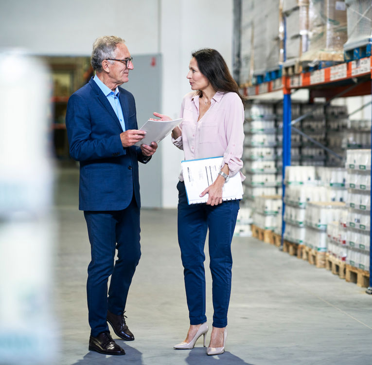 To forretningsfolk diskuterer logistikk på et lager mens de holder dokumenter og et nettbrett.