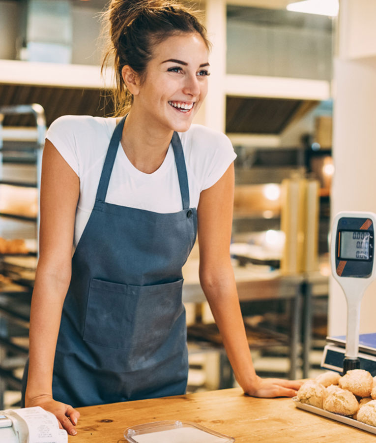 Smilende bakeriansatt med forkle står bak disken med rundstykker og digital vekt – representerer detaljhandel, vaktplanlegging og kundekontakt.
