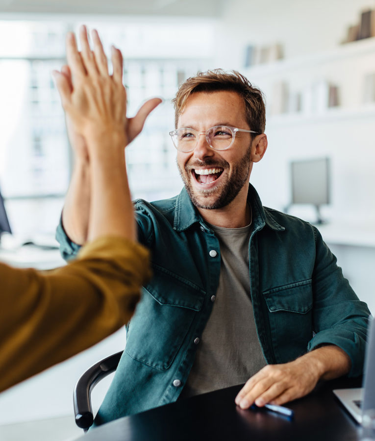 Et entusiastisk team i et moderne kontor feirer en suksess mens en mann og en kvinne gir hverandre en high-five. En annen kollega smiler mens hun jobber på laptopen sin, noe som skaper en positiv og samarbeidsorientert arbeidskultur.