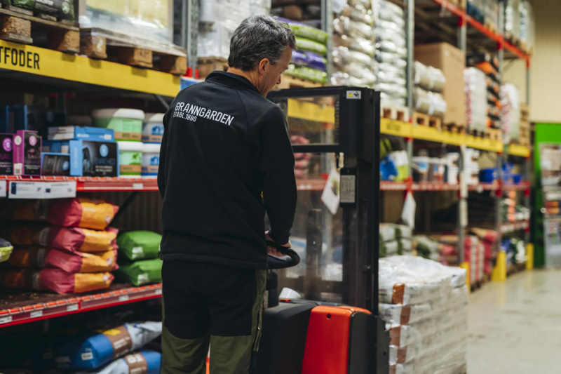 Lagerarbeider hos Grangården håndterer varer med elektrisk palletruck i et godt organisert lager, optimalisert med effektiv vaktplanlegging og timeregistrering.