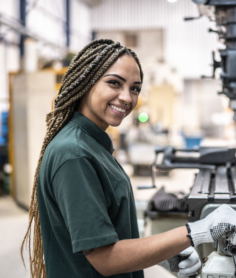 En smilende kvinnelig maskinarbeider i mørkegrønn uniform og hansker bruker en fresemaskin i et produksjonsverksted.