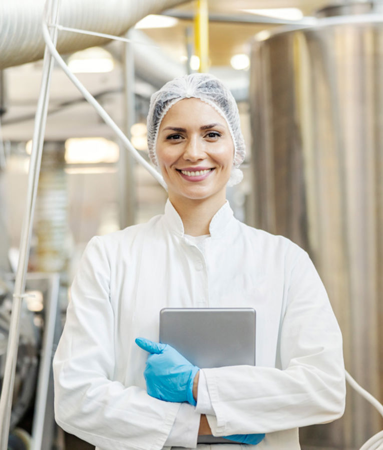 En smilende kvinnelig kvalitetskontrollspesialist i hvit labfrakk, hårnett og hansker holder et nettbrett i en moderne matproduksjonsfabrikk.