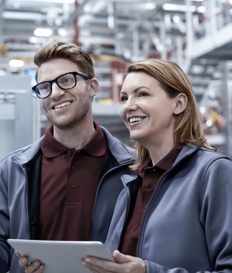 To smilende fabrikkarbeidere bruker et nettbrett og overvåker et kontrollpanel i en avansert produksjonsfasilitet. Representerer arbeidsstyrkeautomatisering og industriell effektivitet.