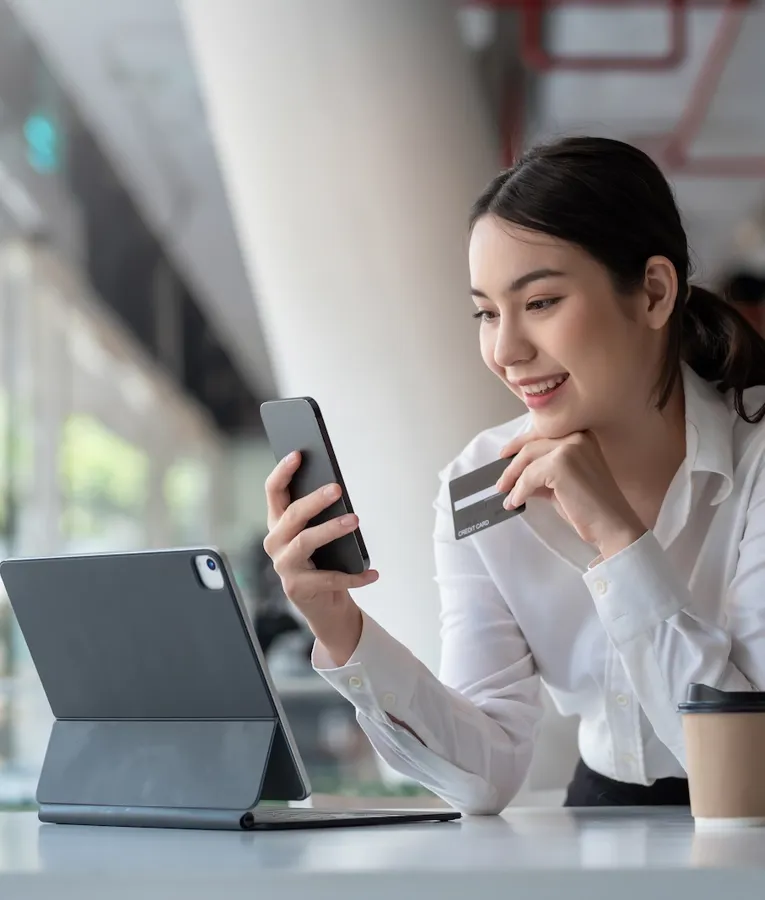 Smilende forretningskvinne bruker en smarttelefon og kredittkort til en nettransaksjon, med et nettbrett og kaffe på bordet, som viser sikker digital betaling.