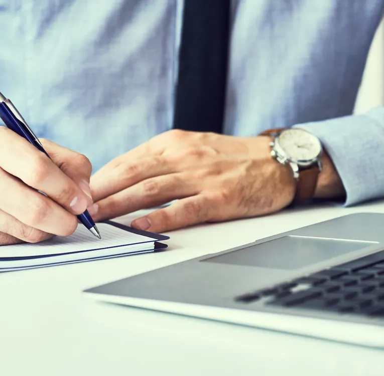 Et nærbilde av en forretningsmann i en blå skjorte og slips som skriver notater i en notatbok mens han jobber på en bærbar PC ved et moderne skrivebord.