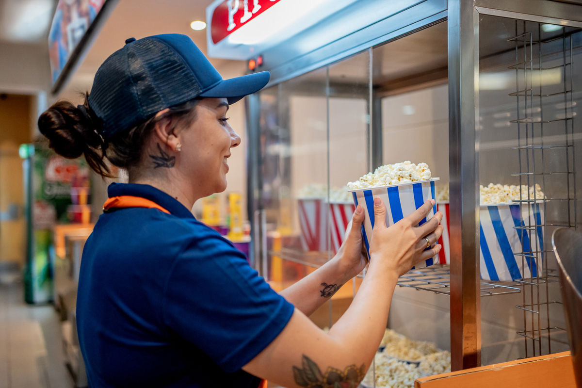 Smilende kinomedarbeider i uniform setter en stripet popcornbøtte i varmeskap – representerer skiftarbeid, kundeservice og effektivt frontpersonell.