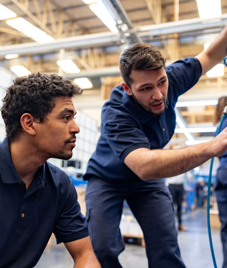 To teknikere i marineblå arbeidsklær samarbeider om utstyrsvedlikehold i fabrikk – illustrerer fagkompetanse, opplæring og teknisk drift.