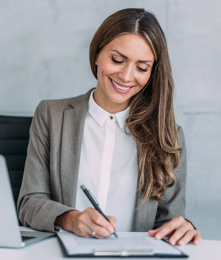 En smilende forretningskvinne i grå blazer signerer dokumenter ved skrivebordet sitt med en bærbar PC ved siden av.