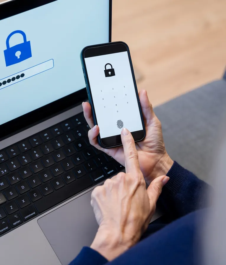 En person bruker biometrisk autentisering på en smarttelefon mens de logger inn på en bærbar PC med en passordskjerm som viser et sikkerhetslåssymbol.