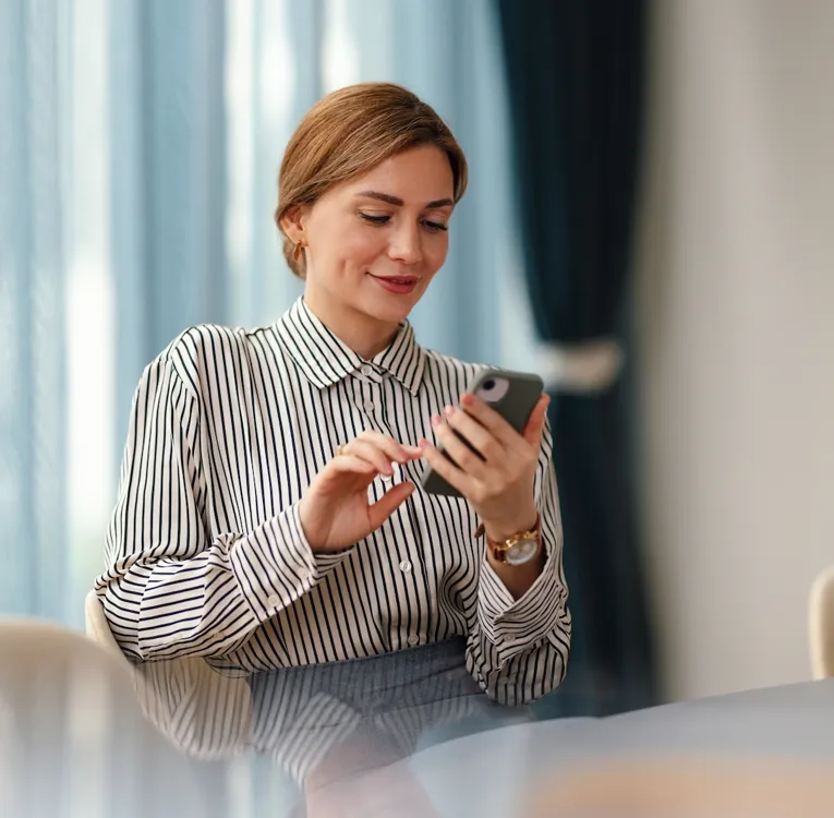 En profesjonell kvinne i en stripete bluse sitter ved et konferansebord og smiler mens hun bruker smarttelefonen sin i et moderne kontor med blå gardiner.