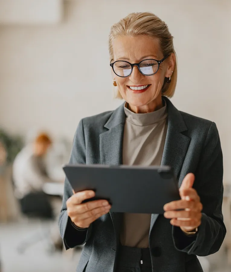 En smilende eldre forretningskvinne i grå dress og briller holder et nettbrett mens hun jobber i et moderne kontor. Kollegaer samarbeider i bakgrunnen, noe som gjenspeiler en produktiv og dynamisk arbeidsplass.