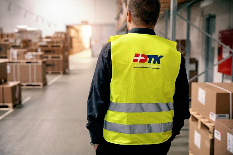 Ansatt i lagerbygning med refleksvest fra DTK overvåker logistikk og bemanning, støttet av effektiv vaktplan og timeregistrering.