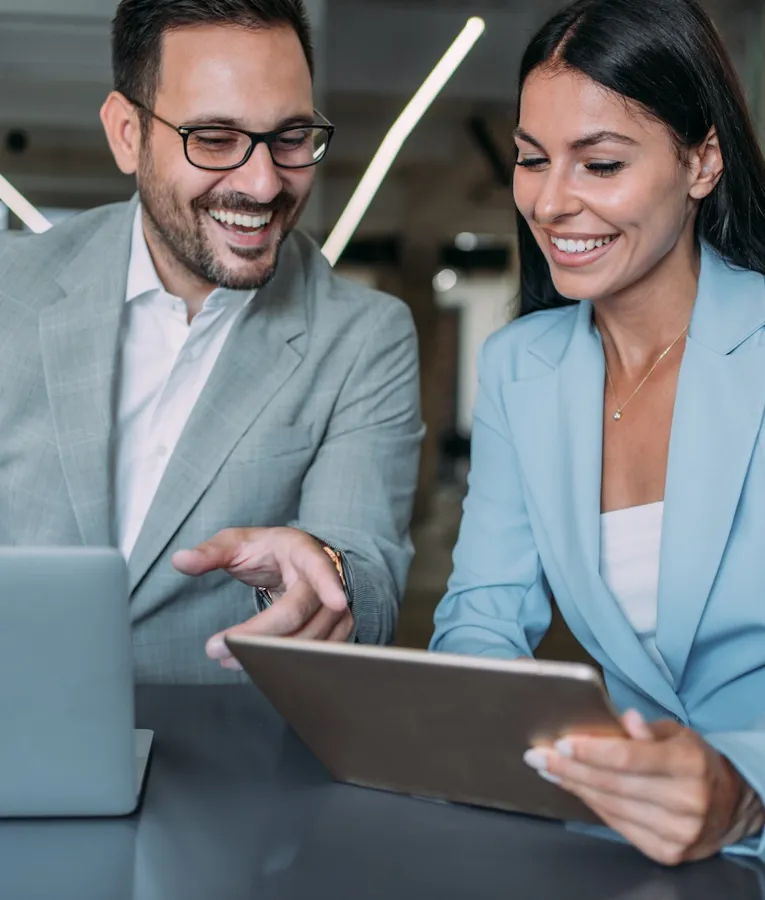 En smilende forretningskvinne i en lyseblå blazer holder et nettbrett mens hun samarbeider med en mannlig kollega i grå dress, som peker på en laptop-skjerm. De er i et moderne kontor og diskuterer arbeidsstyring.