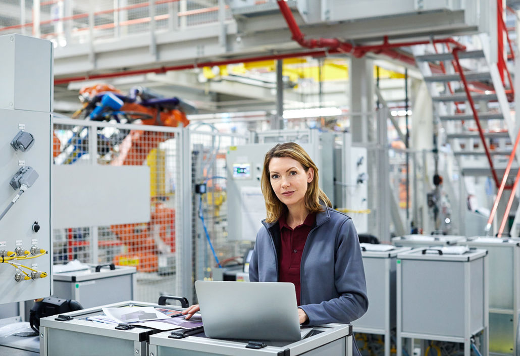 En selvsikker kvinnelig ingeniør jobber på en bærbar PC i en moderne industrifabrikk og overvåker automatiserte produksjonssystemer.