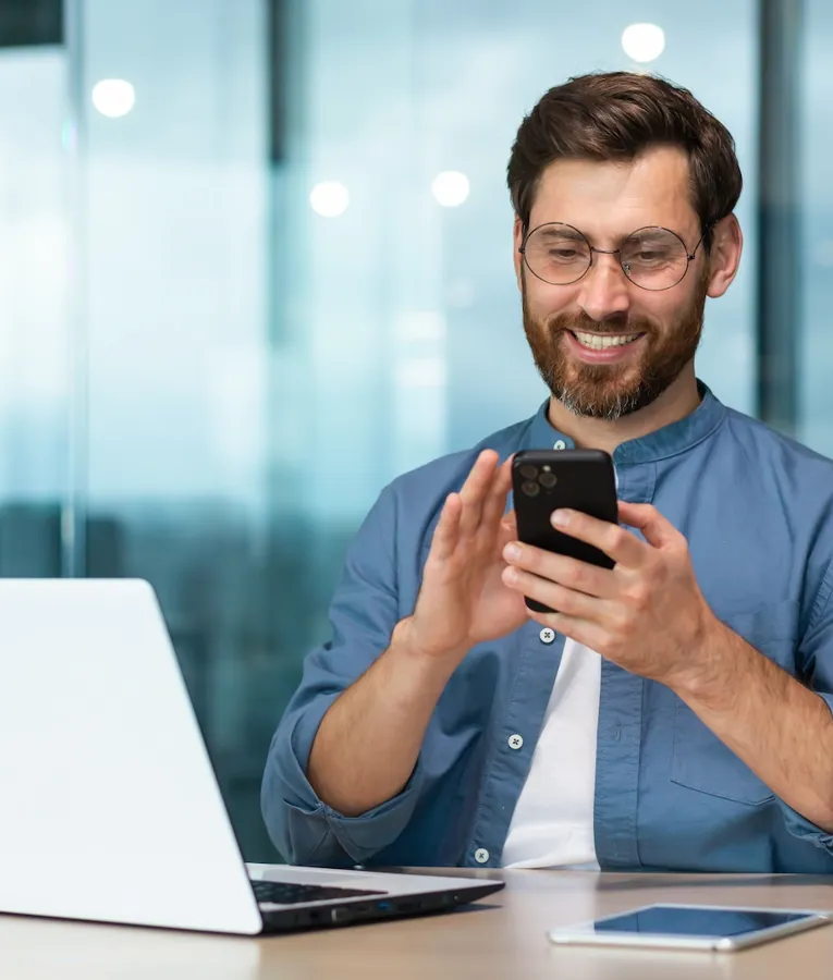 Smilende forretningsmann bruker en smarttelefon ved skrivebordet sitt