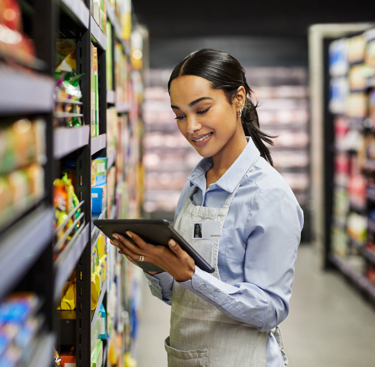 En smilende supermarkedansatt i en blå skjorte og forkle bruker et digitalt nettbrett for å sjekke varelageret mens hun jobber i butikkens gang.