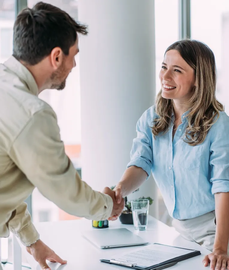 Smilende kollegaer håndhilser over et skrivebord i et lyst kontormiljø – representerer vellykket samarbeid, onboarding og god bedriftskultur.