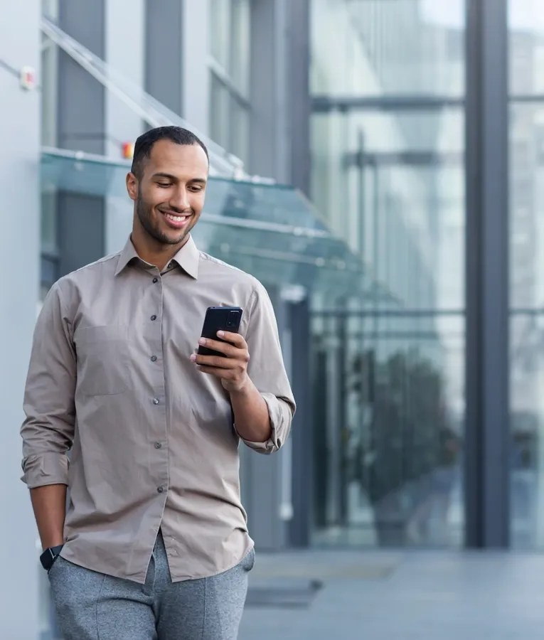 En smilende profesjonell i en beige skjorte går utenfor en moderne kontorbygning i glass mens han sjekker smarttelefonen sin, som illustrerer en mobil og fleksibel arbeidshverdag.