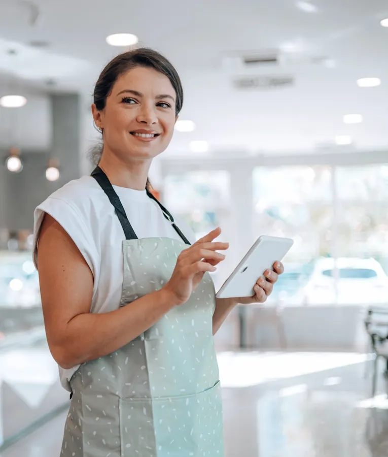 Smilende bakeriansatt bruker et nettbrett til lager- eller ordrebehandling i en moderne kafé, som viser digitale arbeidsstyringsløsninger.