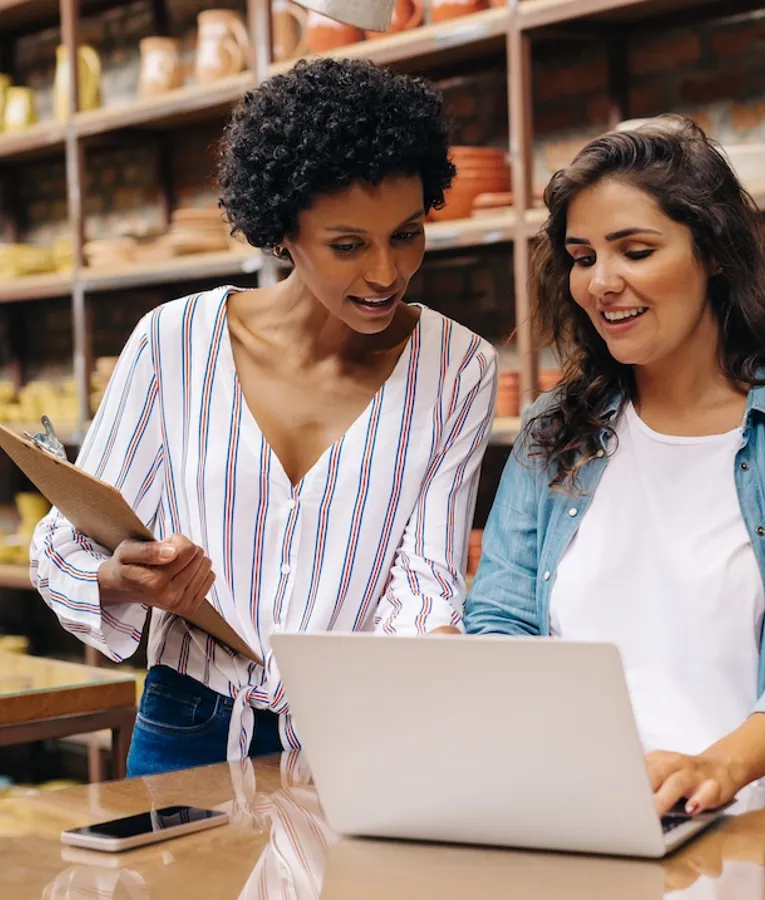 To smilende butikkansatte jobber sammen ved en butikkdisk, bruker en bærbar PC for å administrere lager og salg, omgitt av keramikkprodukter.