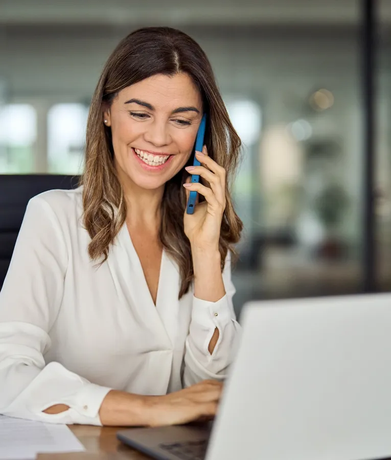 Smilende forretningskvinne snakker i telefon mens hun jobber på en bærbar PC i et moderne kontor, som viser effektiv kommunikasjon for fjernarbeid.